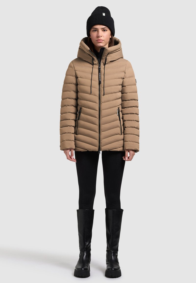 Frau in beiger Pufferjacke mit Kapuze, schwarzem Beanie, schwarzen Leggings und schwarzen kniehohen Stiefeln, die vor einem schlichten Hintergrund steht.
