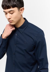 Camisa de botón azul marino en una mezcla de algodón, con un cuello clásico y un sutil bordado de logo en el pecho. Textura suave.
