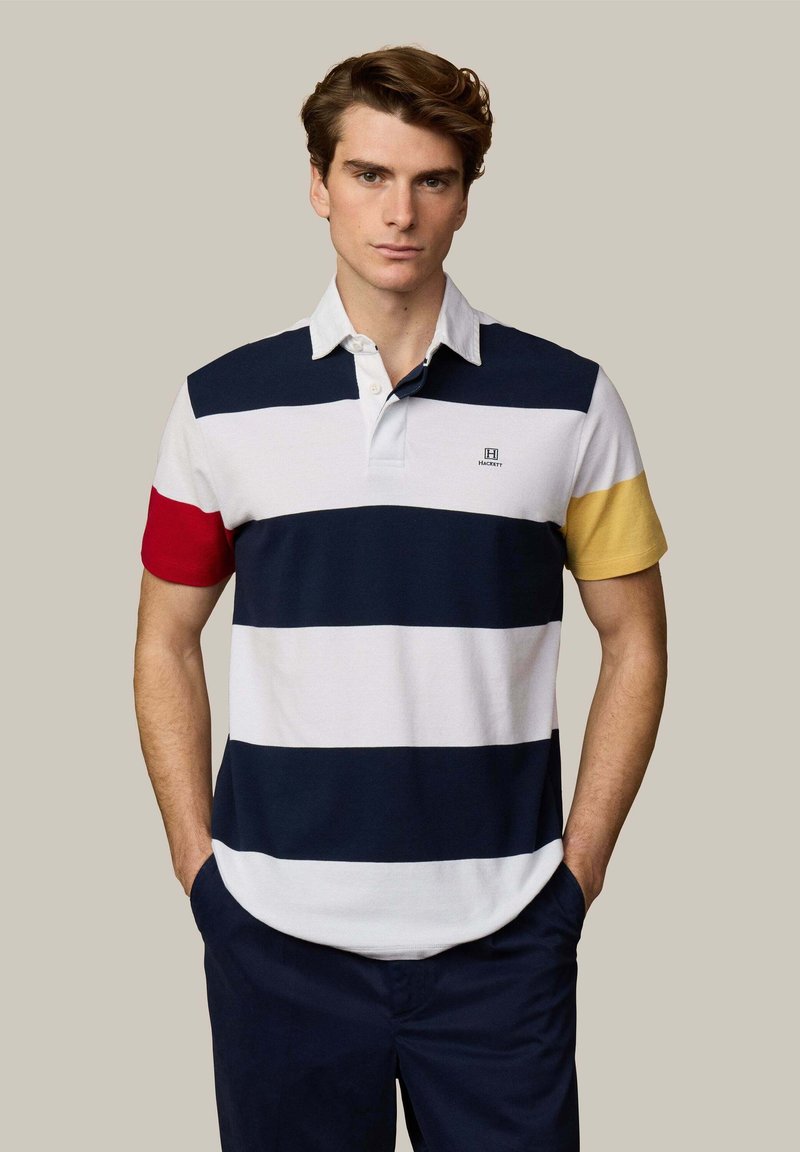 Homme les mains dans les poches portant un polo à manches courtes rayé bleu marine, blanc, rouge et jaune, ainsi qu'un pantalon bleu marine, sur un fond uni.