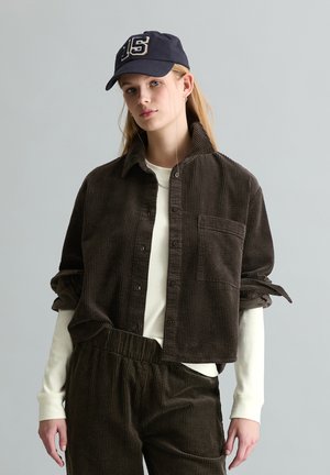 Młoda kobieta ubrana w ciemnobrązową kurtkę i spodnie z sztruksu, kremową koszulę z długim rękawem oraz granatową czapkę z numerem 15, stojąca na jednolitym szarym tle.