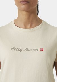 Krémové tričko s krátkým rukávem s nápisem "Helly-Hansen" v kurzívě, s malým akcentem norské vlajky. Jemný na dotek.