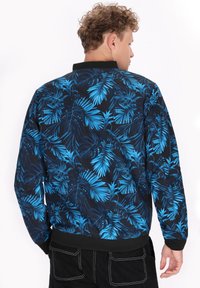 Blauwe en zwarte bomberjack met een tropisch bladpatroon. Beschikt over een geribde kraag en manchetten, een gladde textuur en een elastische tailleband.