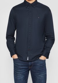 Man som bär en mörk marinblå knäppt skjorta med en liten logotyp på bröstet och blå jeans, händerna i fickorna.