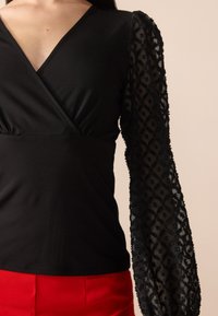Haut noir avec un décolleté en V profond, corps ajusté et manches longues transparentes présentant un motif floral texturé. Associé à un pantalon rouge taille haute.