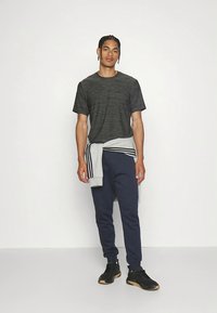 T-shirt desportivo preta e cinza com um design texturizado, combinada com calças de ganga azul-marinho. Uma sweatshirt cinza clara amarrada à cintura. Ténis pretos.