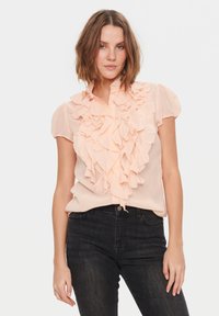 Blusa color pesca con dettagli arricciati sul fronte, maniche corte a sbuffo e colletto con bottoni. Abbinata a jeans neri aderenti.