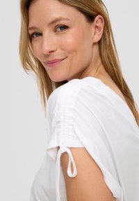 Top bianco con maniche arricciate e dettaglio con cordoncino. Tessuto morbido, vestibilità rilassata, con una leggera drappeggiatura intorno alle spalle.