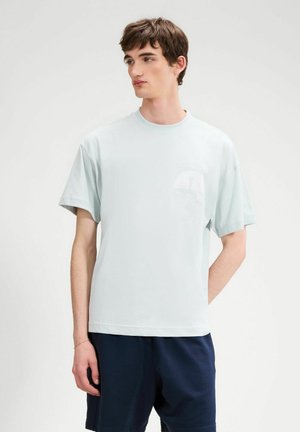 Junger Mann trägt ein hellblaues, übergroßes T-Shirt mit weißem Logo und marineblaue Shorts, steht vor einem schlichten weißen Hintergrund.