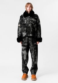 Chaqueta de patrón negro y gris con cuello de piel sintética, combinada con pantalones holgados a juego, complementada con calzado naranja.