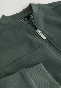 Dunkelgrüne, strukturierte Jacke mit silbernem Reißverschluss, geripptem Kragen und Bündchen, auf einem hellen Hintergrund flach ausgelegt.