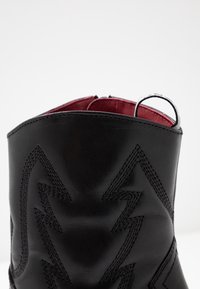 Gros plan sur une botte en cuir noir avec des coutures en zigzag et une doublure intérieure rouge, dotée d'une languette au sommet.