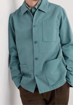 Mand iført teal farvet jakke med knapper og bryst- samt sidelommer over en ternet skjorte og brune bukser, hænderne i lommerne.