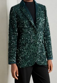 Mörkgrön paljettblazer med en satinliknande krage, två framknappar och en texturerad yta. Bärs över en svart topp och matchas med svarta byxor.