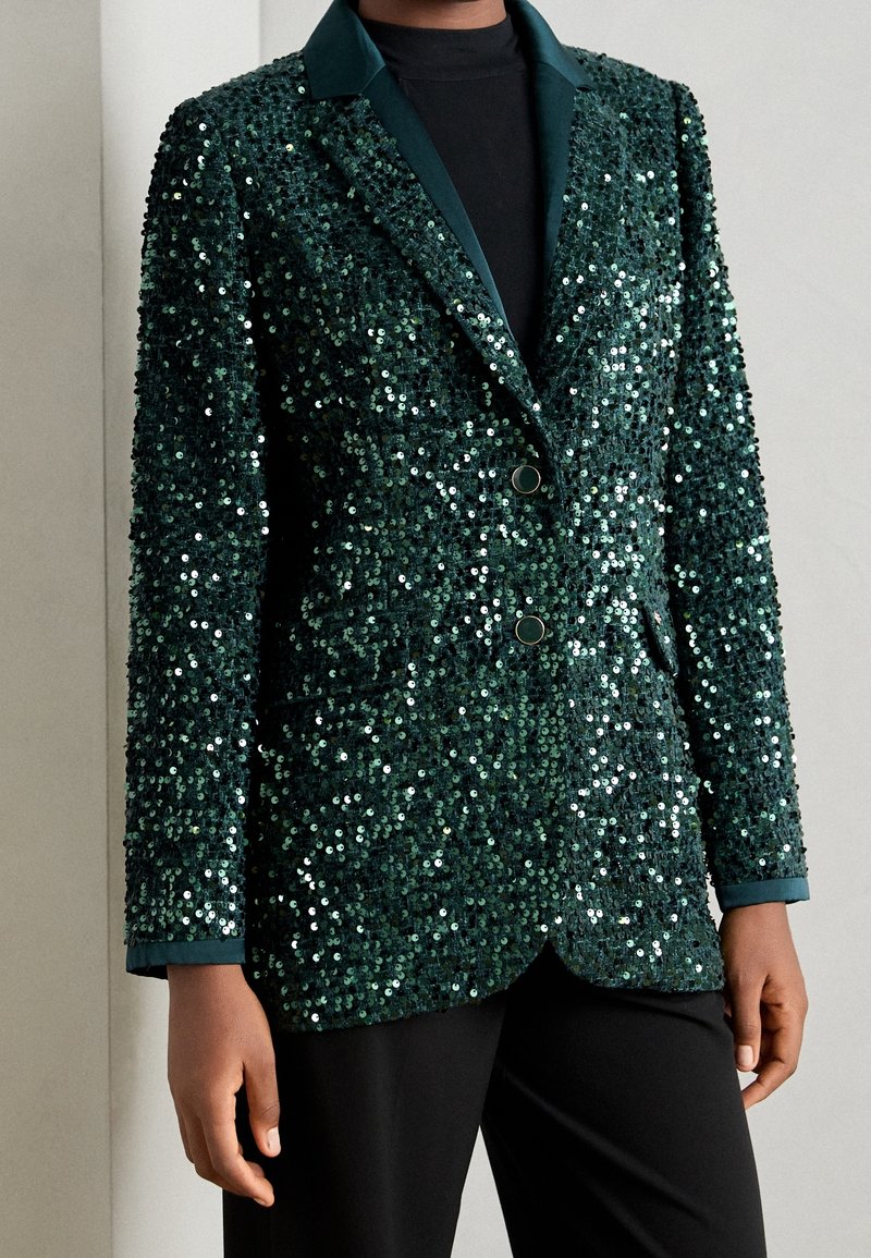 Mörkgrön paljettblazer med en satinliknande krage, två framknappar och en texturerad yta. Bärs över en svart topp och matchas med svarta byxor.