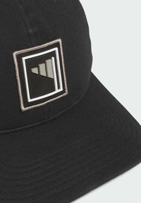 Černá čepice s texturovanou tkaninou, zdobená obdélníkovou vyšívanou nášivkou zobrazující geometrické tvary v šedé a bílé barvě.