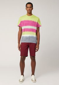 Camiseta de manga corta en patrón tie-dye con rayas verdes, rosas y grises, combinada con pantalones cortos color burdeos y zapatillas blancas.