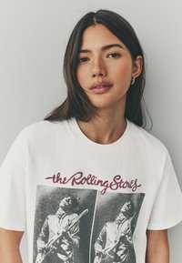 Hvid bomulds t-shirt med et grafisk print af to musikere, sort-hvide billeder, og "the Rolling Stones" i mørkerød skrift.