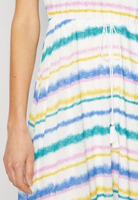 Main reposant à côté d'une robe blanche à rayures horizontales rose, jaune, bleue et bleu sarcelle, avec une ceinture à nouer ornée de pampilles.