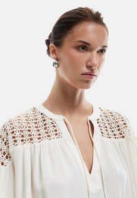 Blusa bianca con dettagli in pizzo sulle spalle, tessuto strutturato e scollatura a V. Modella che indossa orecchini metallici, capelli raccolti in uno chignon.