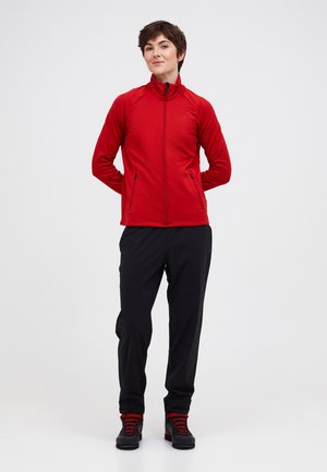 Jeune personne debout, mains derrière le dos, portant une veste zippée rouge, un pantalon noir et des chaussures de randonnée noires avec des lacets rouges.