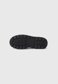 Semelle de chaussure noire avec un motif de semelle chunky et des rainures profondes pour l'adhérence, présentée à plat sur un fond clair.