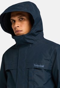 Navy blauwe waterdichte jas met capuchon, voorzien van een ritssluiting aan de voorkant, twee borstzakken en het Timberland-logo in lichtblauwe borduring.