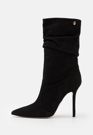 Musta mokkanahkainen nilkkuri, jossa on terävä kärki ja korkea tikkikorko, löysä muotoilu ja kullanvärinen yksityiskohta yläosassa.