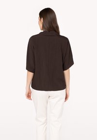 Blusa marrone a maniche corte in tessuto morbido e testurizzato, con cucitura sul retro e una vestibilità rilassata, abbinata a pantaloni bianchi.