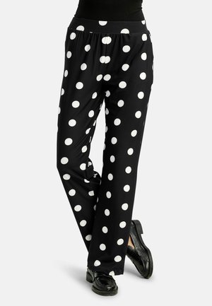 Čierne nohavice s širokými nohavicami a veľkými bielymi bodkami, nosené s čiernymi mokasínmi, nohy skrížené v členkoch.