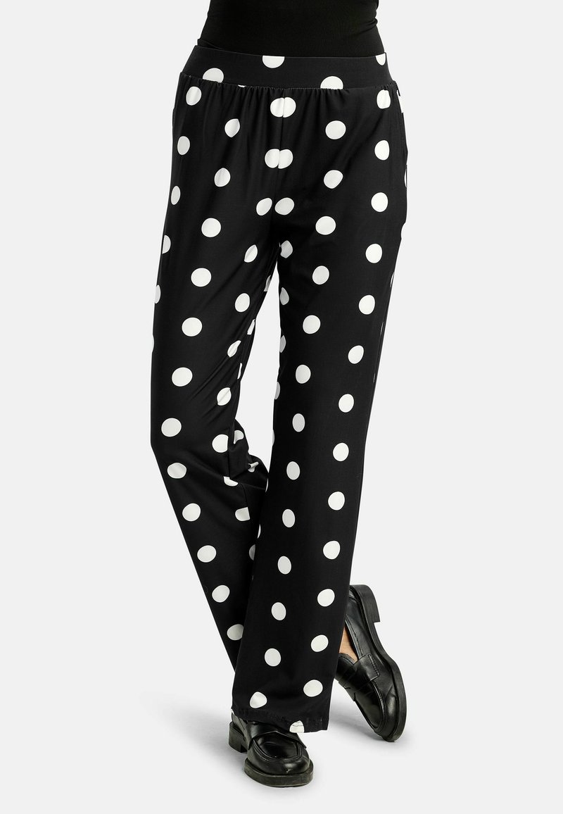 Juodos plačios kelnės su didelėmis baltomis taškuotėmis, dėvimos su juodais loaferiais, kojos sukryžiuotos ties čiurnomis.