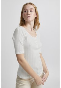 Hvid kortærmet bluse med rund halsudskæring, der har en knaplukning og et tekstureret mønster af små diamanter over stoffet.