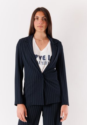 Donna che indossa un abito blu navy a righe sopra una camicia bianca con il testo parzialmente visibile "Love is Beautiful", in piedi davanti a uno sfondo semplice.