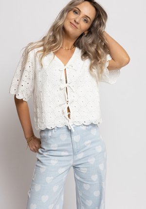 Vrouw met golvend blond haar draagt een witte blouse met oogjes en een strik aan de voorkant, en lichtblauwe broek met een patroon van witte hartjes, poseert met één hand in het haar.