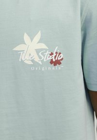 Camiseta de algodón azul clara con mangas cortas, que presenta gráficos florales y el texto "The Studio Originals" en blanco.