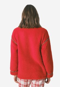 Czerwony sweter z polaru z fakturowaną powierzchnią, długimi rękawami i dekoracyjnym wykończeniem przy ramionach oraz mankietach. Pokazano widok z tyłu.