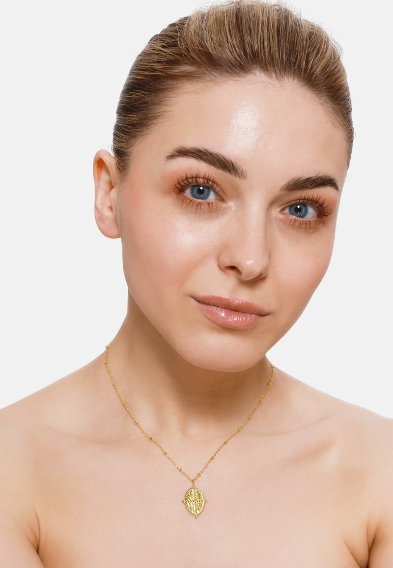 Collana d'oro con un pendente a forma di foglia decorata, caratterizzata da dettagli intricati e una catena delicata, indossata su pelle nuda.