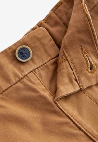 Gros plan sur la ceinture d'un pantalon marron avec un bouton à quatre trous cousu, montrant les coutures détaillées et la boutonnière.