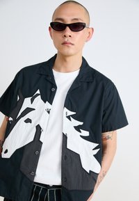 Short-sleeve black shirt featuring a white horse head appliqué. Material is lightweight, with buttons and a casual collar design.