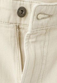 Gros plan sur un jean en denim beige montrant un bouton métallique estampillé « MNG DENIM » et une fermeture éclair argentée partiellement ouverte.