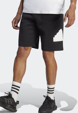 Mężczyzna w czarnych szortach Adidas z dużym białym logo, białych skarpetach w czarne paski i czarnych butach sportowych Adidas, stojący na jasnym tle.