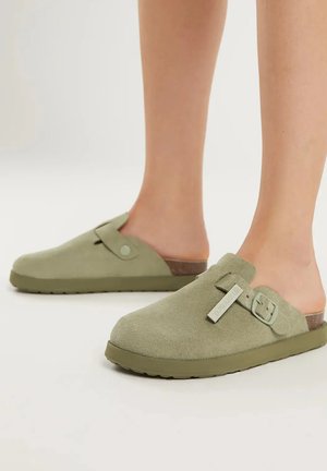 Personne portant des sabots en daim vert olive à enfiler avec des brides à boucles réglables, debout sur un fond clair uni.