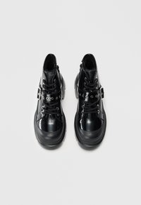 Et par glinsende svarte lær ankelboots med tykke såler, snøring og et dekorativt belte med metalløyler og spenne på hvit bakgrunn.