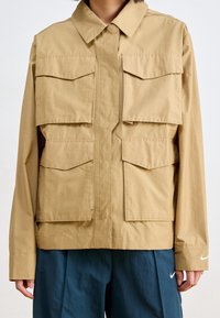 Beżowa, lekka kurtka z zapięciem na guziki z przodu, dużymi kieszeniami na piersiach i bokach oraz klasycznym kołnierzem. Na rękawie znajduje się logo Nike.