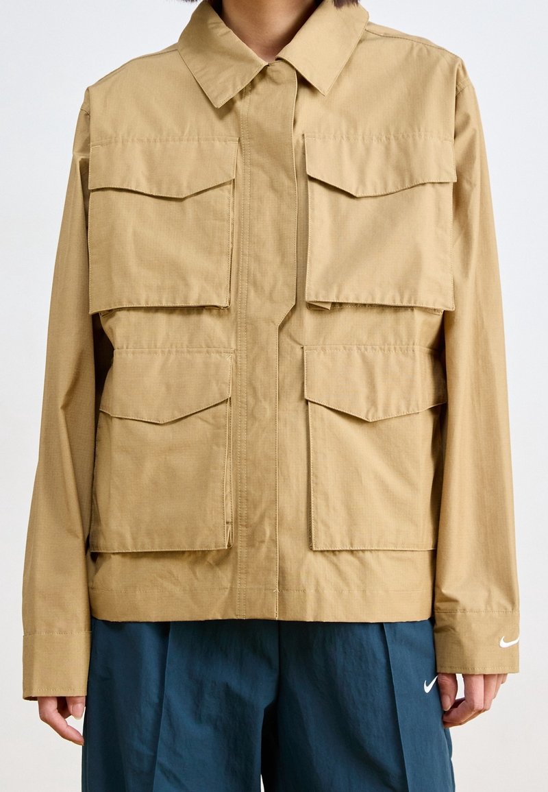 Beżowa, lekka kurtka z zapięciem na guziki z przodu, dużymi kieszeniami na piersiach i bokach oraz klasycznym kołnierzem. Na rękawie znajduje się logo Nike.