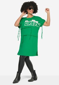 Vestido camiseta oversized de algodón verde con texto "GREEN" en blanco, aberturas laterales y detalles de cordón. Combinado con leggings brillantes negros y botas con tachuelas.