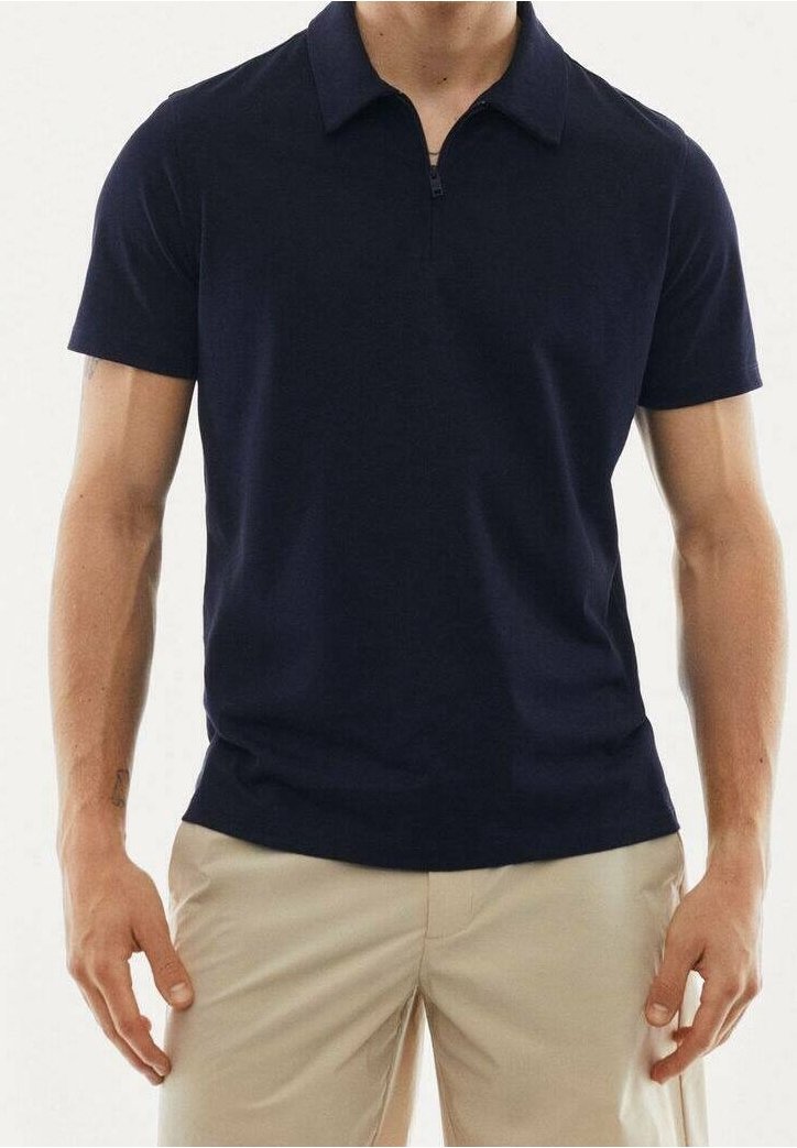 Homme portant un polo bleu marine à manches courtes avec col zippé et pantalon beige, debout devant un fond uni.