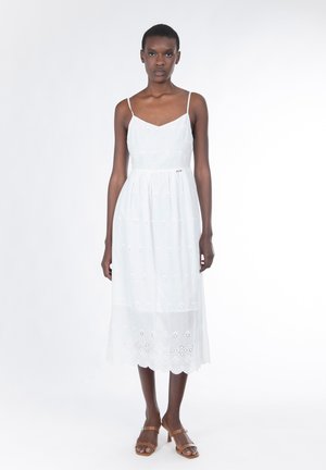 Weißes, ärmelloses Kleid mit eng anliegendem Oberteil und ausgestelltem Saum mit Spitzenbesatz. Mit floralen Lochstickmustern und dünnen Trägern. Getragen mit braunen Sandalen.