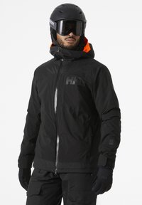 Veste de ski noire avec fermeture éclair à l'avant, col haut et accents orange. Présente un logo sur la poitrine et des poignets élastiques. Portée avec un casque noir et des lunettes de protection.