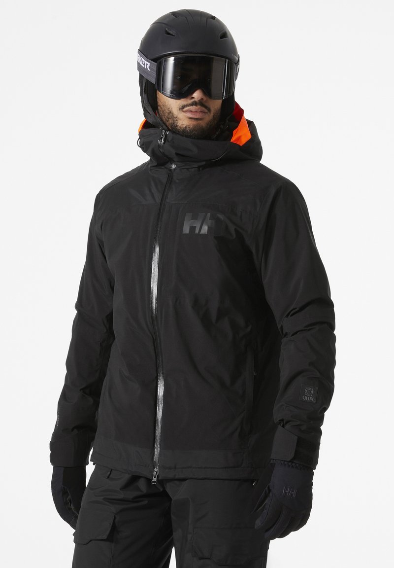 Veste de ski noire avec fermeture éclair à l'avant, col haut et accents orange. Présente un logo sur la poitrine et des poignets élastiques. Portée avec un casque noir et des lunettes de protection.