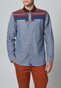 Camisa de manga larga azul claro con cuello marrón oscuro, con canesú estampado en rosa y azul, y dos bolsillos en el pecho.